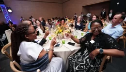 People at a formal dinner toast, raising wine glasses around a long table with flowers and candles.
